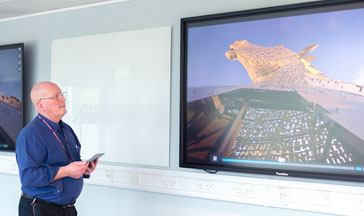 A  staff member standing by 2 screens showing the Scottish Kelpies sculpture