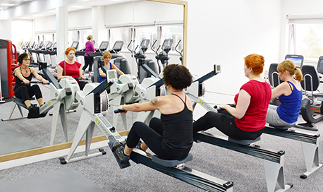 A row of women using rowing machines in front of a mirrored wall, 