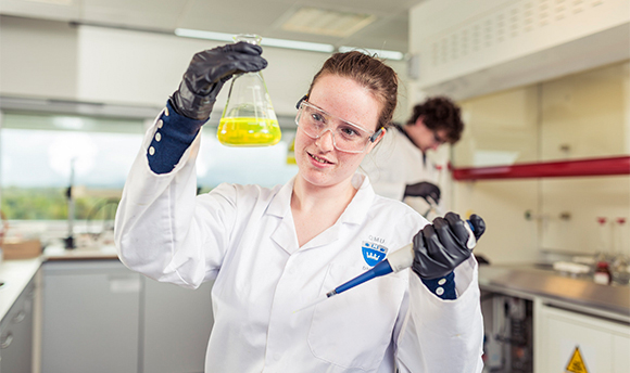 A  student wearing PPE and holding up a beaker of yellow liquid